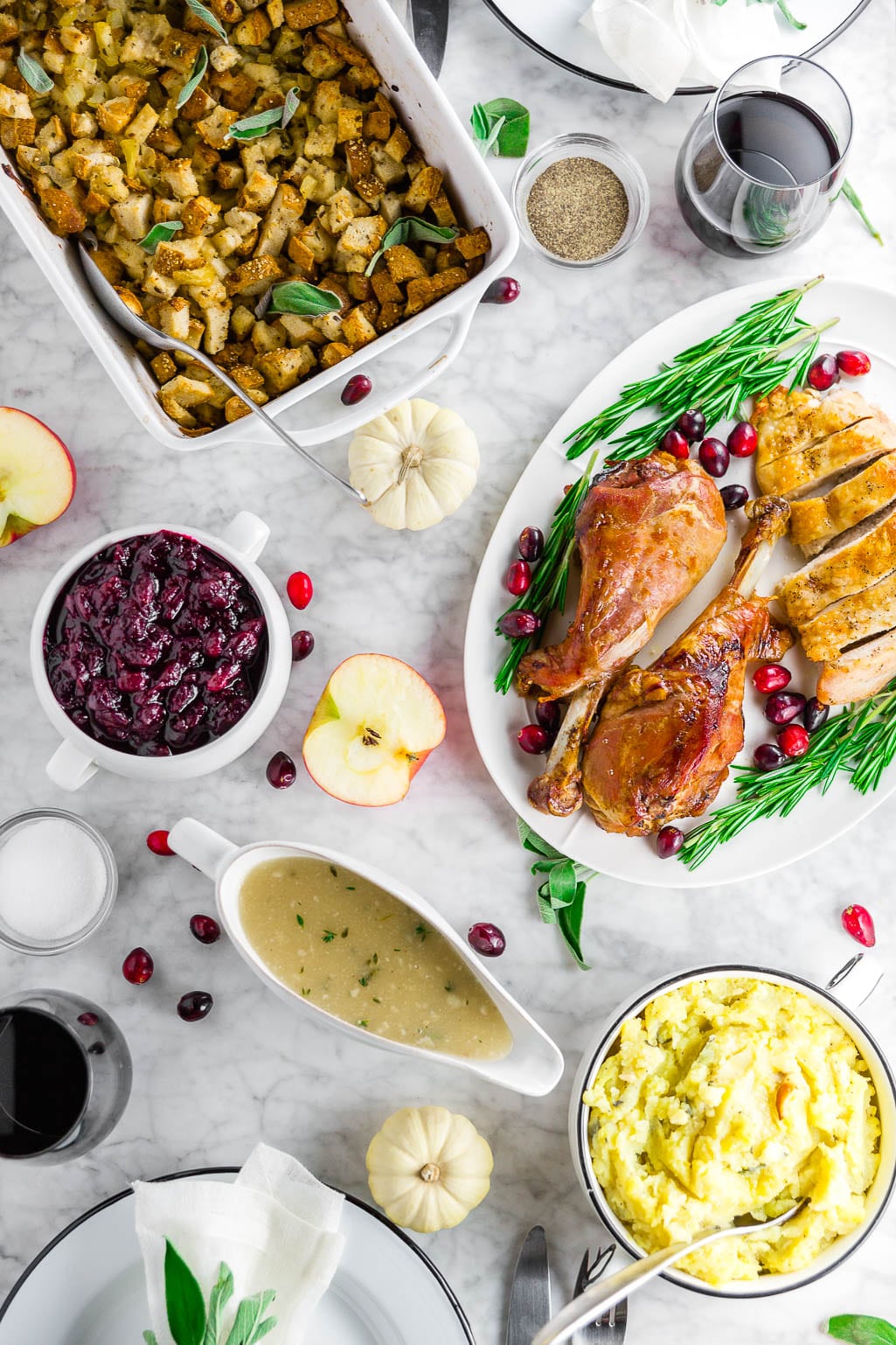 A white marble table with a Thanksgiving feast with gluten-free stuffing, turkey legs with cranberries and rosemary, cranberry sauce, gluten-free grave, a bowl of mashed potatoes, and two glasses of red wine with place settings. 