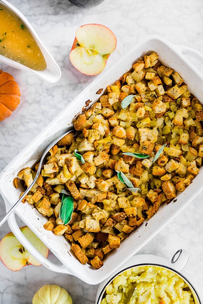 A white baking sheet with classic gluten-free stuffing with celery, onion and apple with a bowl of mashed potatoes and gravy. 