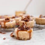 A mini pecan pie cheesecake with pecan pie caramel sauce dripping down the sides and a cooling rack with pecan pie cheesecakes in the background.