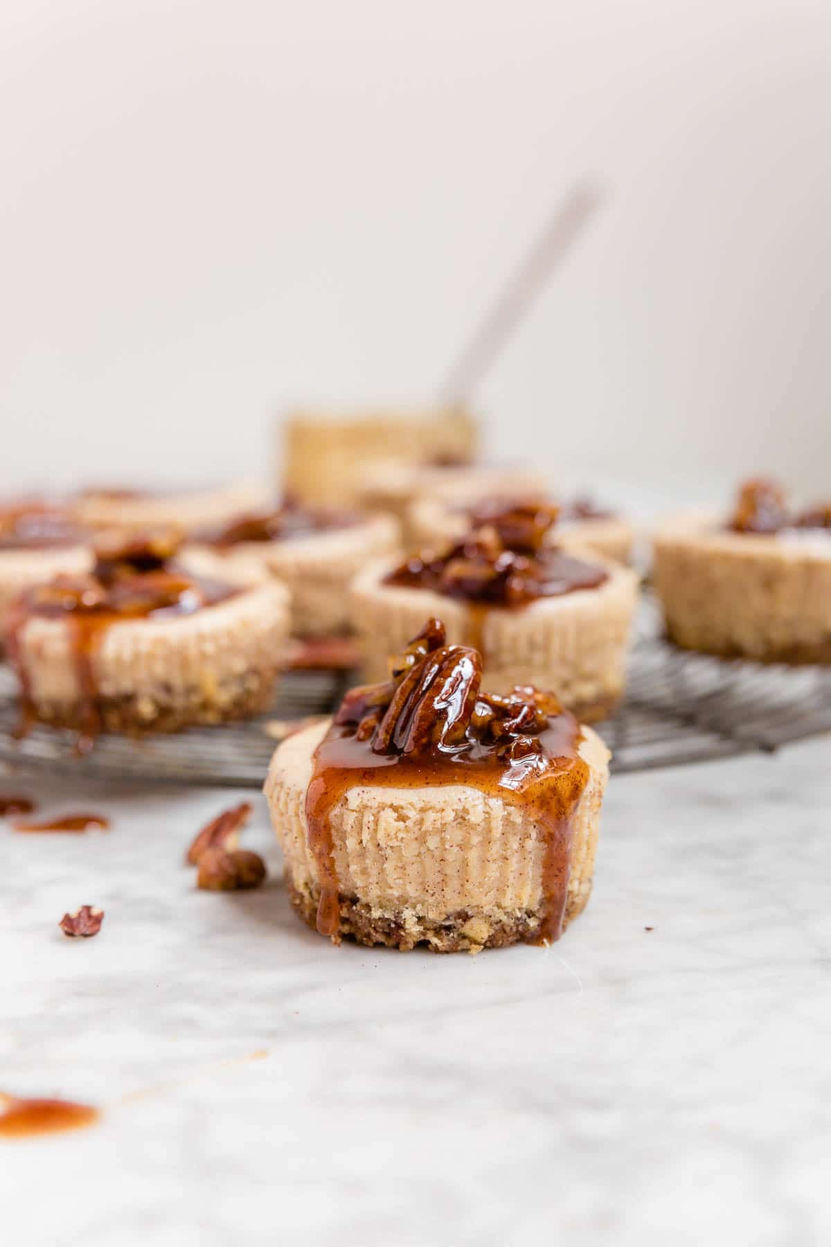 A mini pecan pie cheesecake with pecan pie caramel sauce dripping down the sides and a cooling rack with pecan pie cheesecakes in the background. 