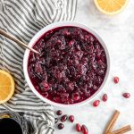 A bowl of red wine cranberry sauce with two orange halves, cinnamon sticks, fresh cranberries and a glass of red wine.