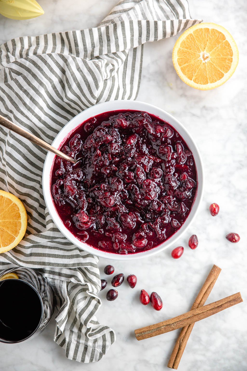 A bowl of red wine cranberry sauce with two orange halves, cinnamon sticks, fresh cranberries and a glass of red wine. 