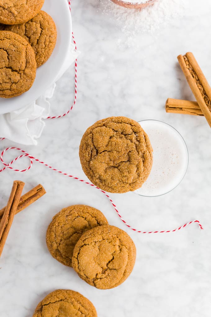 A ginger molasses cookie on top of a glass of milk with a plate of chewy molasses cookies and cinnamon sticks. 