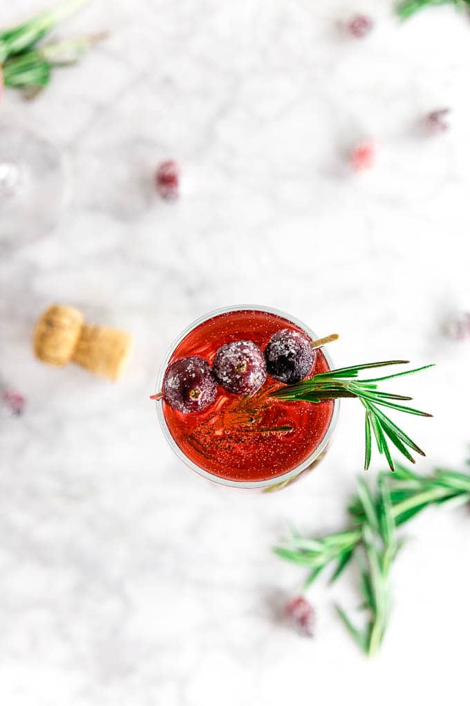 A champagne flute filled with cranberry mimosa topped with fresh rosemary and sugared cranberries on a marble table