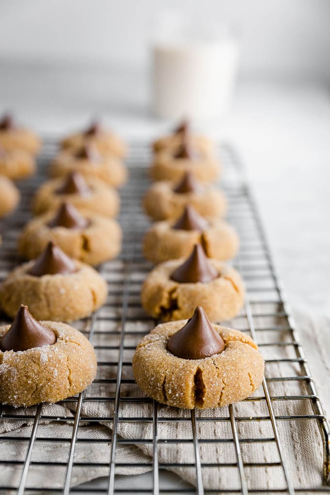 rows of peanut butter blossoms with a chocolate kiss on top on a cooling rack with a glass of milk. 