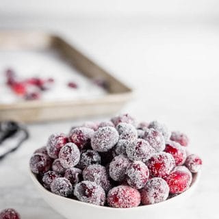A bowl of sugared cranberries with a baking sheet with granulated sugar and fresh cranberries.
