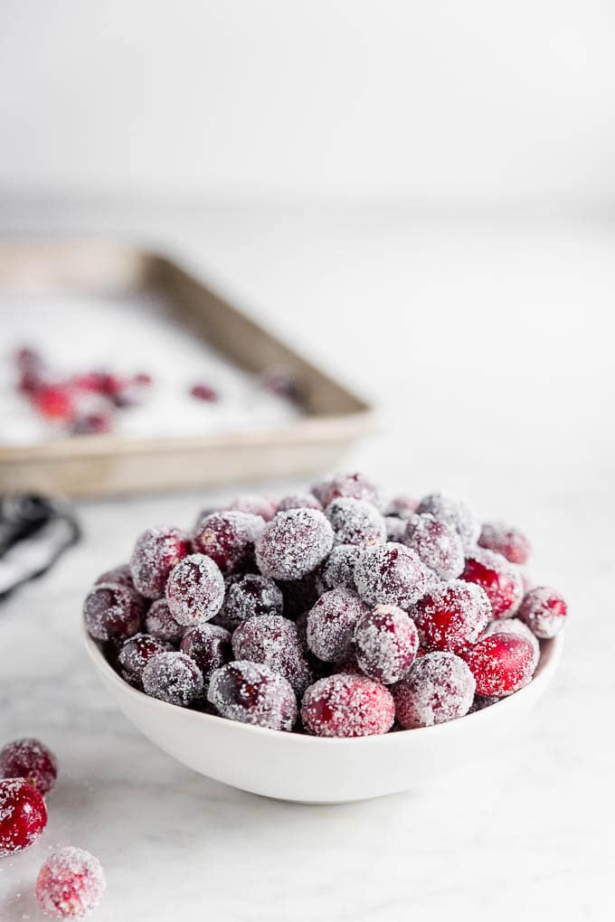 A bowl of sugared cranberries with a baking sheet with granulated sugar and fresh cranberries. 