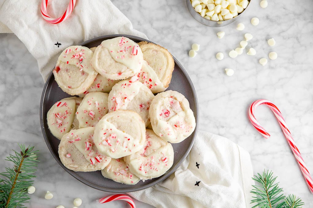 A plate of gluten-free icebox cookies dipped in white chocolate and crushed candy canes with winter greens, candy canes, and a cup of white chocolate chips.