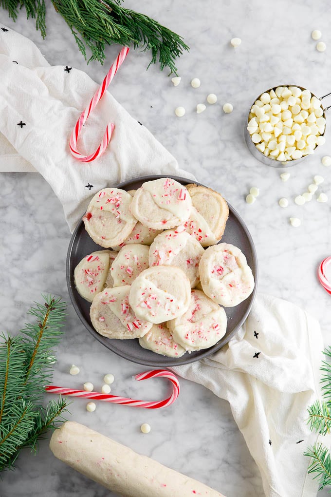 A plate of slice and bake cookies dipped in white chocolate and topped with crushed candy canes surrounded by a log of cookie dough, a measuring cup of white chocolate chips, winter greenery and candy canes. 