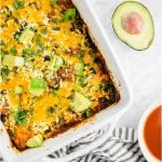 A baking dish filled with gluten-free chicken enchilada casserole with red enchilada sauce, shredded cheddar cheese, diced avocado and cilantro with a bowl of enchilada sauce and an avocado.