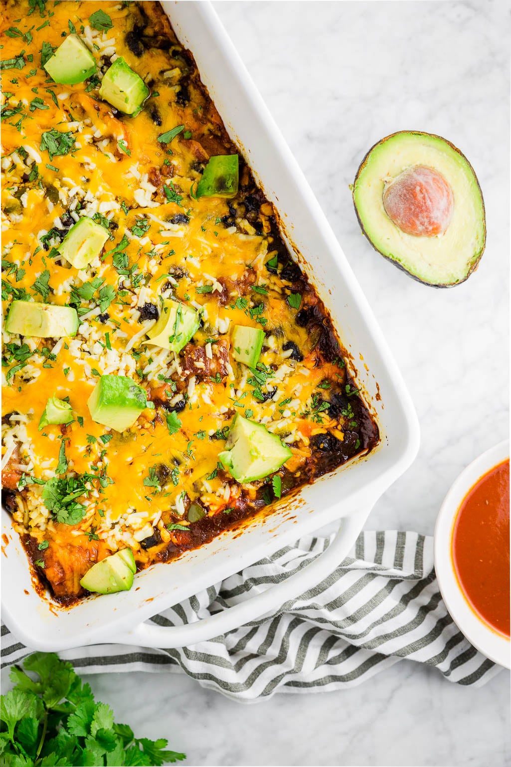 A baking dish filled with gluten-free chicken enchilada casserole with red enchilada sauce, shredded cheddar cheese, diced avocado and cilantro with a bowl of enchilada sauce and an avocado.