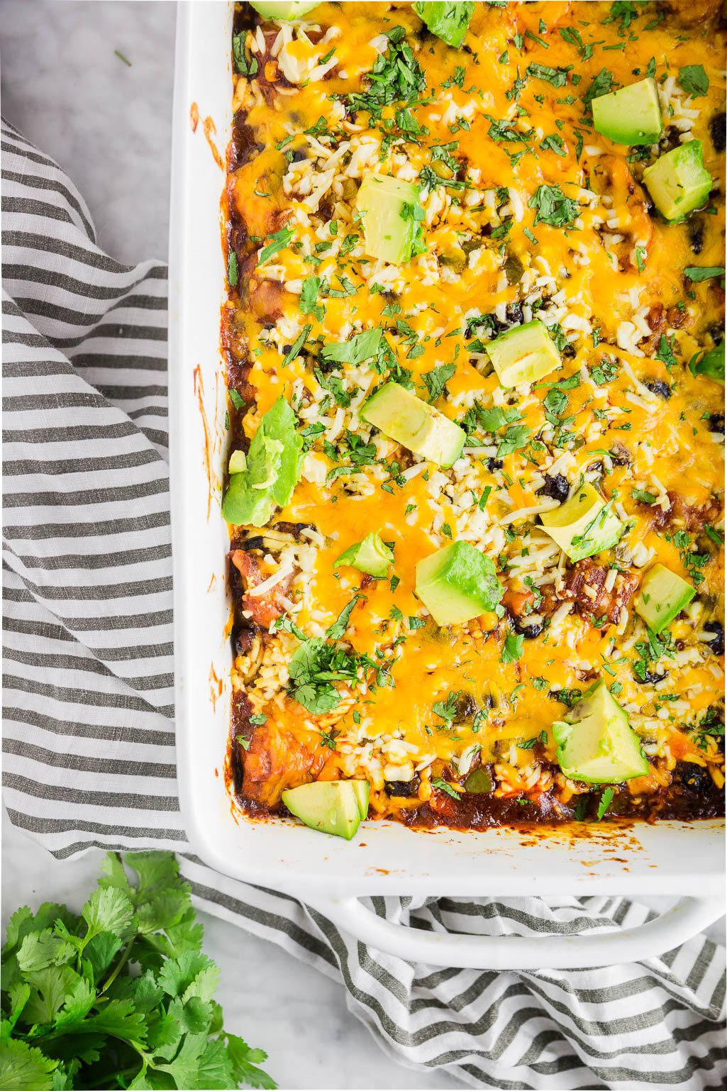 A white baking dish filled with chicken enchilada casserole topped with shredded cheese, avocado slices, and chopped cilantro.