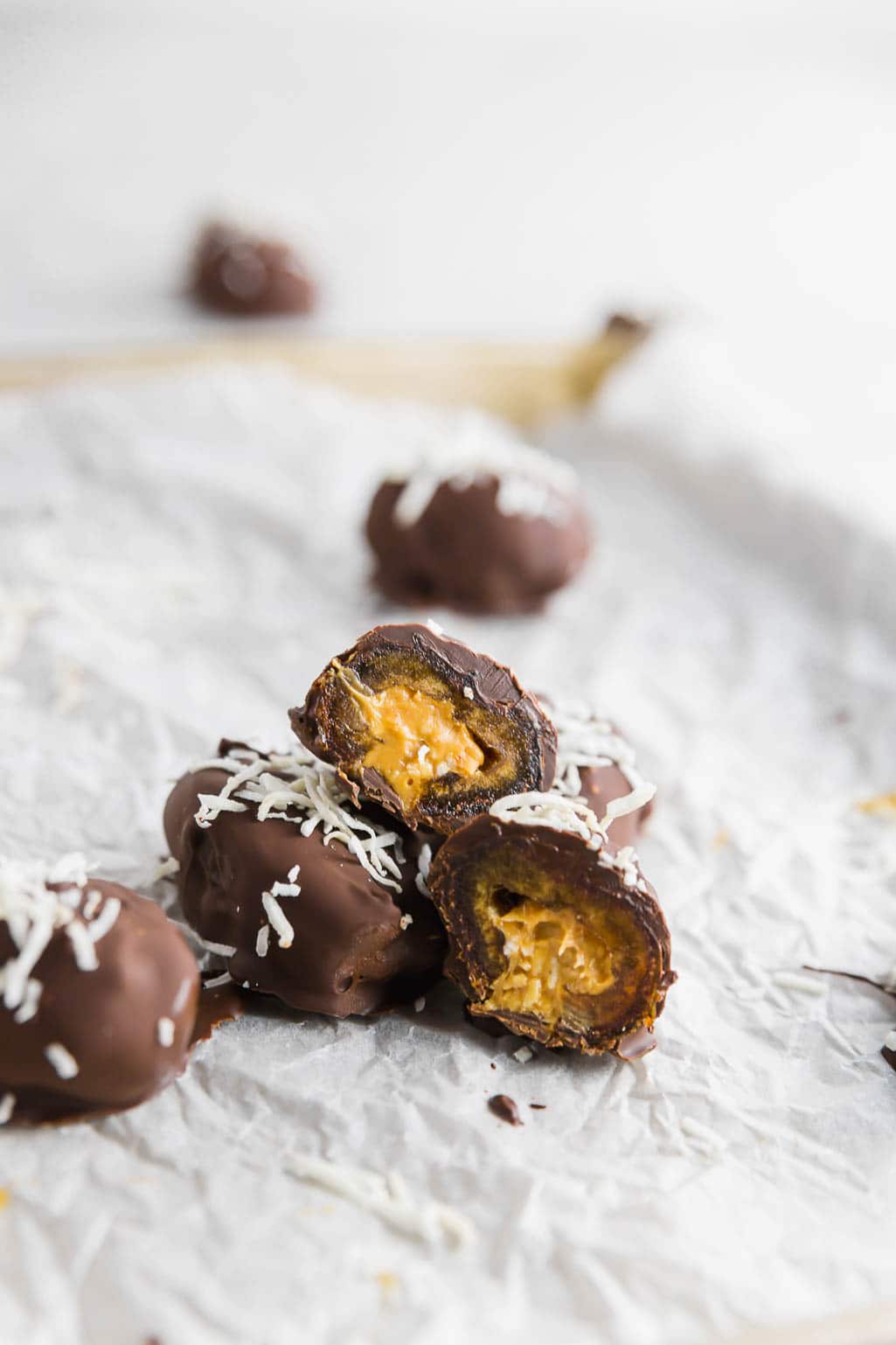 Several chocolate covered peanut butter stuffed dates topped with shredded coconut on white parchment paper. 