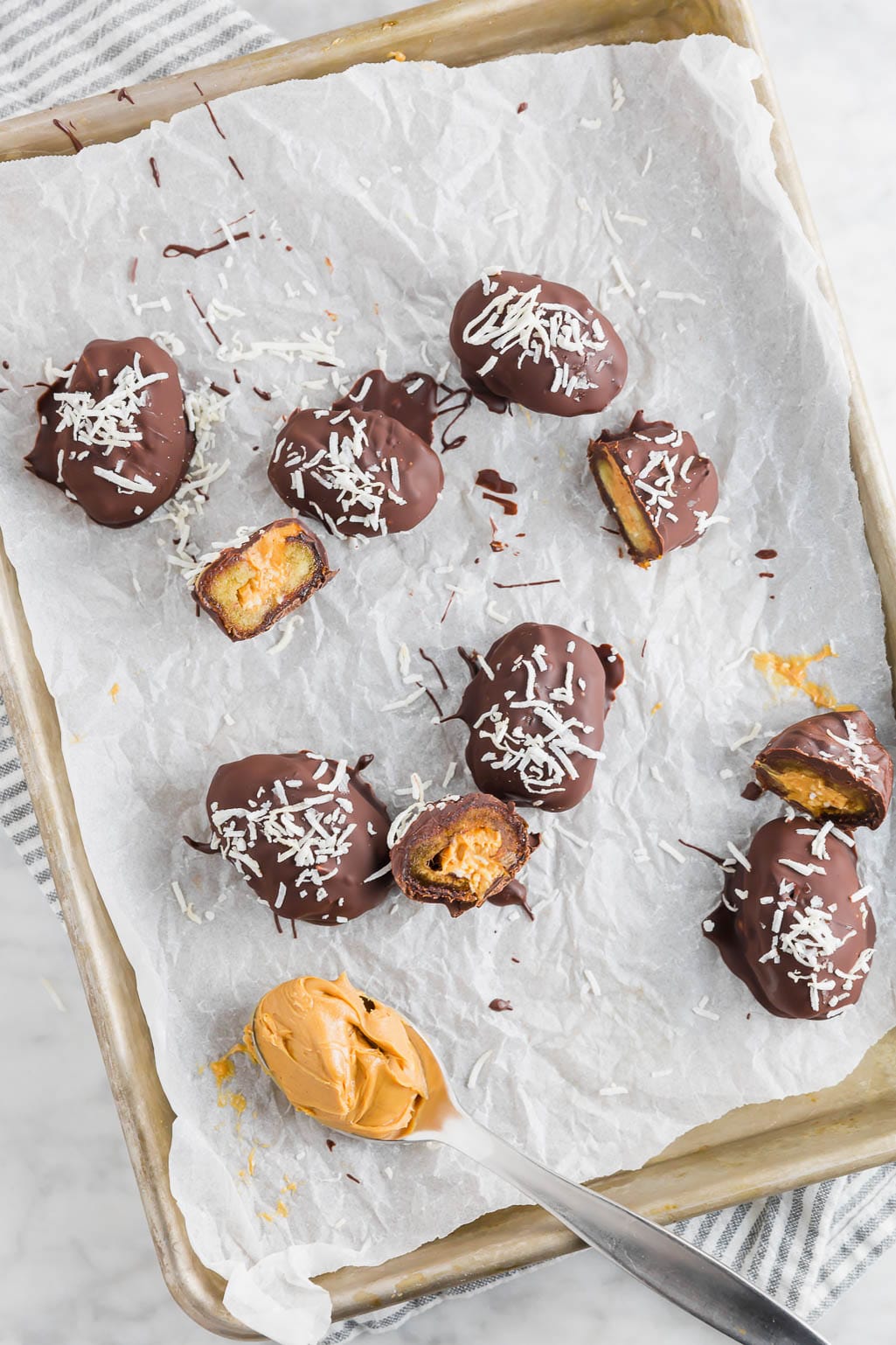A sheet pan lined with parchment paper with a spoonful of peanut butter and chocolate covered peanut butter stuffed dates topped with coconut. 