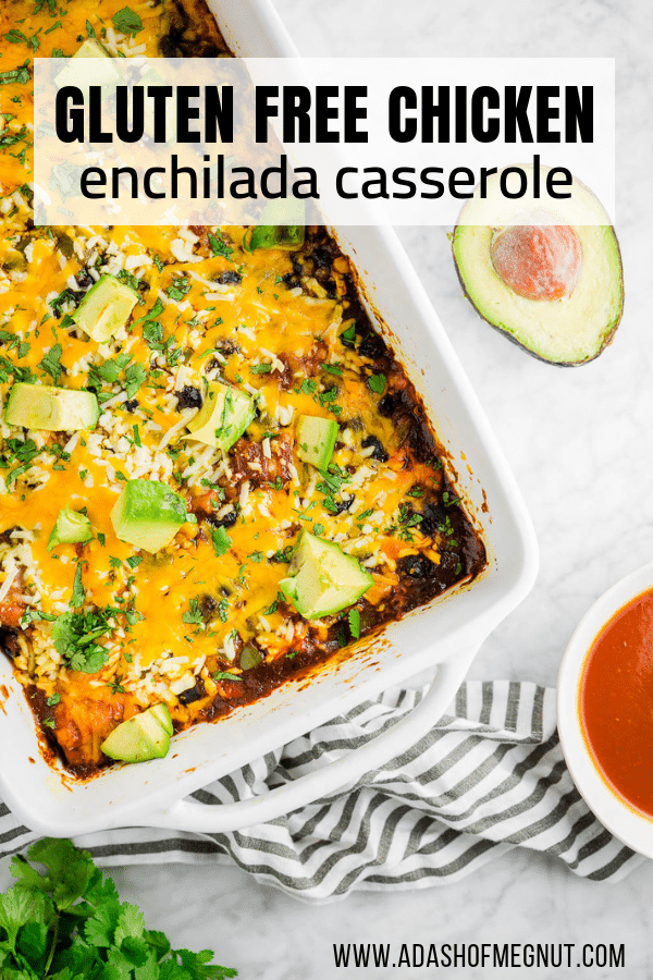 A baking dish filled with gluten-free chicken enchilada casserole with red enchilada sauce, shredded cheddar cheese, diced avocado and cilantro with a bowl of enchilada sauce and an avocado.