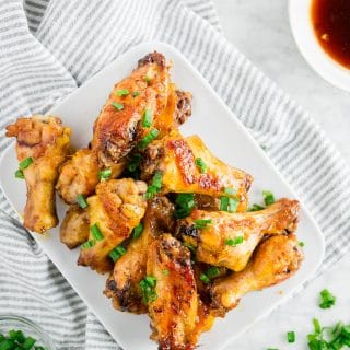 A plate of baked honey garlic chicken wings topped with green onions on a white table with a side of honey garlic sauce.
