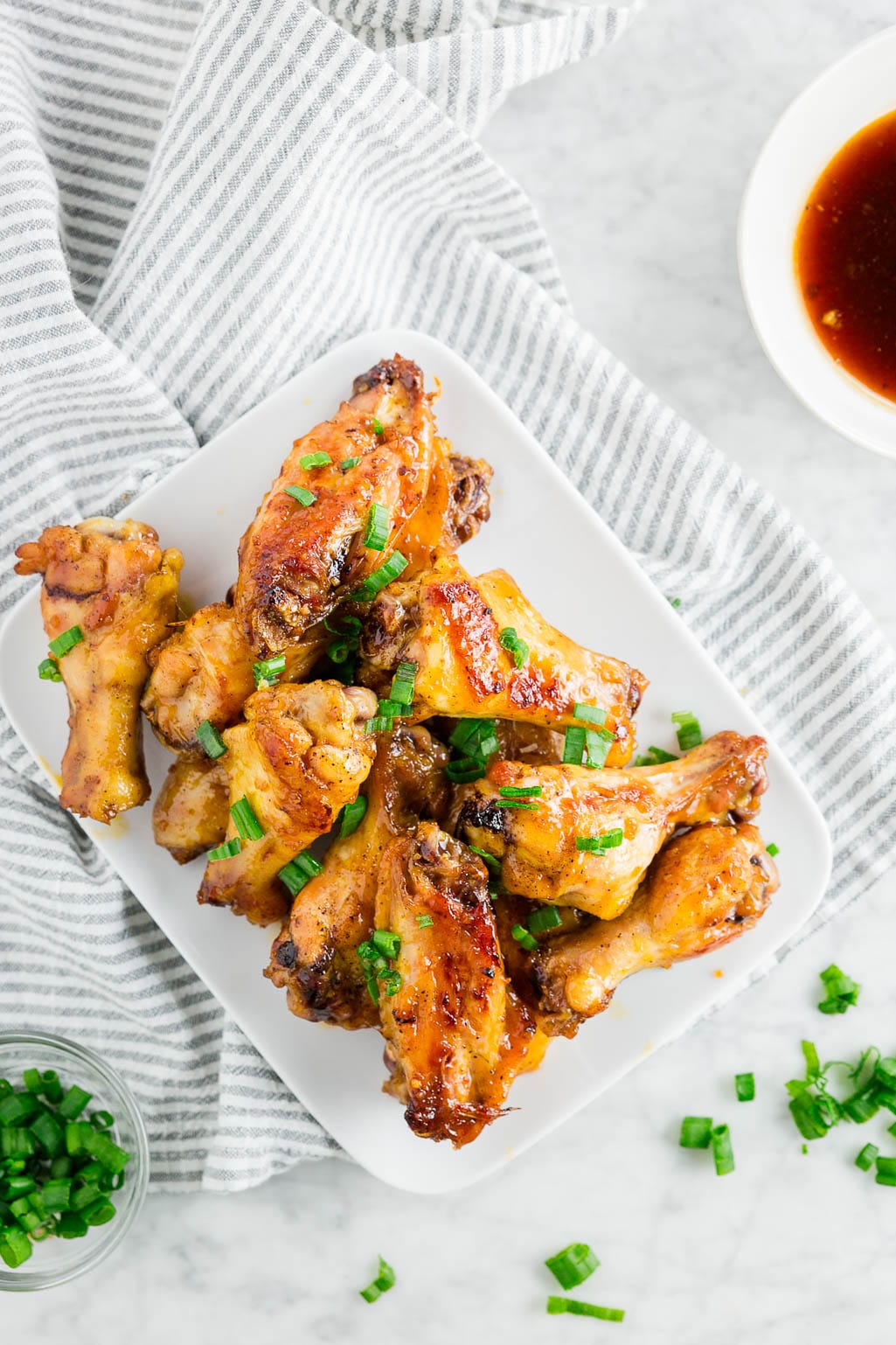 A plate of baked honey garlic chicken wings topped with green onions on a white table with a side of honey garlic sauce.