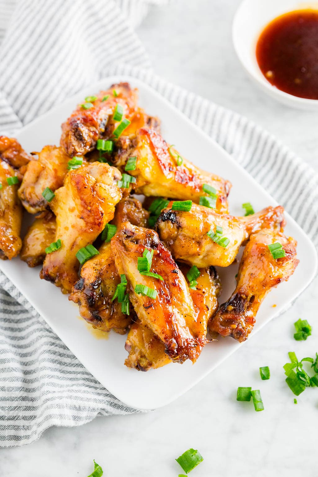 A plate of baked honey garlic chicken wings topped with green onions on a white table with a side of honey garlic sauce.