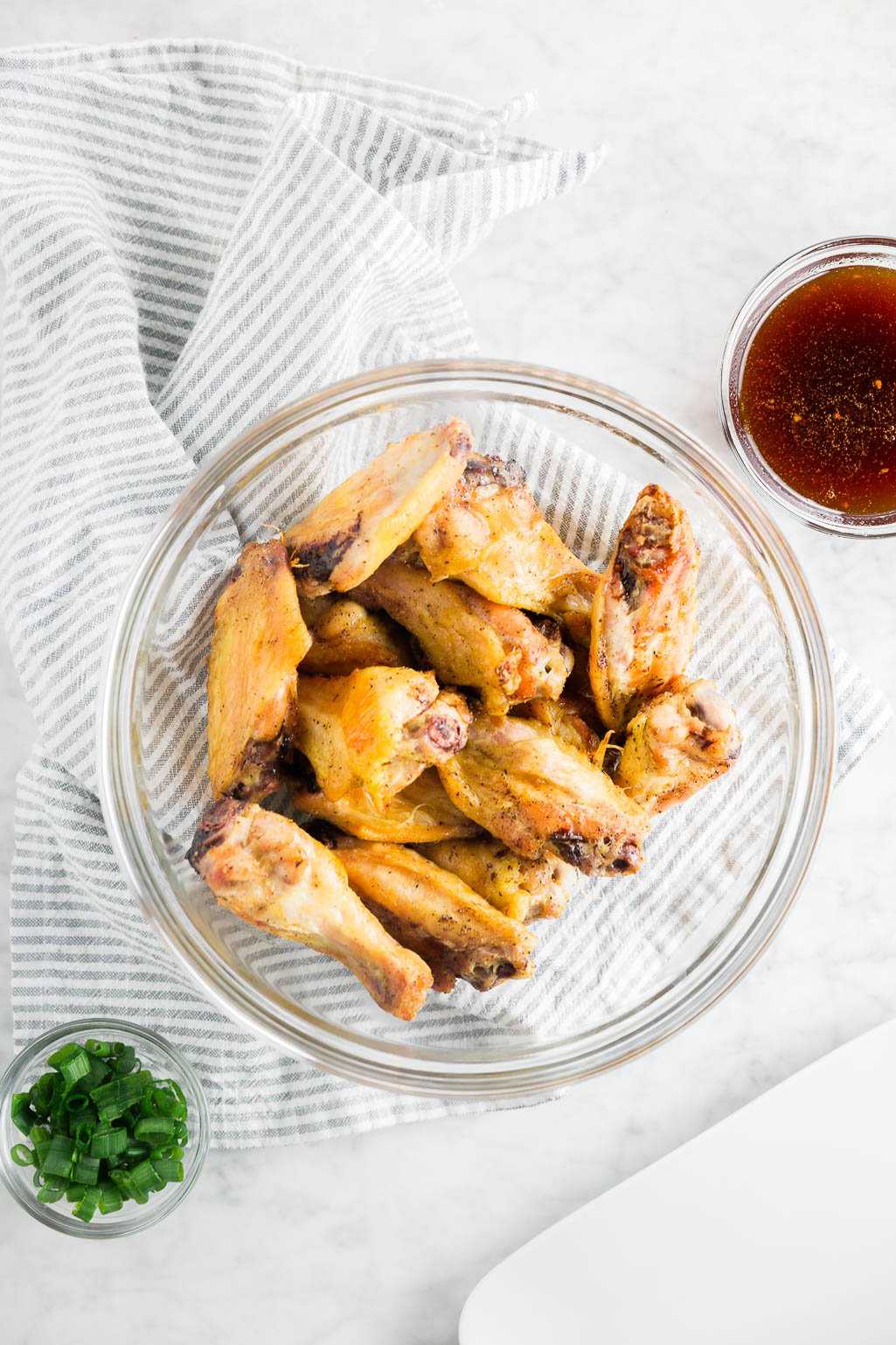 A bowl of baked chicken wings waiting for honey garlic sauce to be poured over the top with a bowl of green onions. 