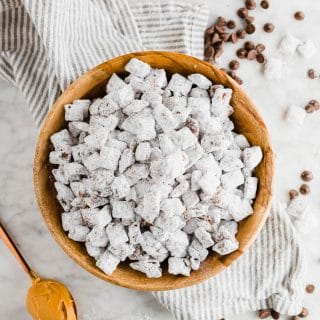 Dairy-free puppy chow in a wooden bowl with a spoonful of peanut butter and chocolate chips and powdered sugar on a linen napkin and table.