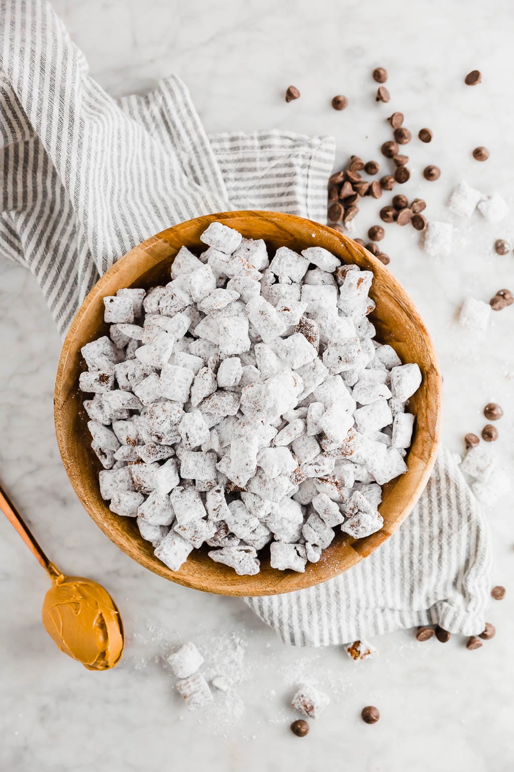 Dairy-free puppy chow in a wooden bowl with a spoonful of peanut butter and chocolate chips and powdered sugar on a linen napkin and table.