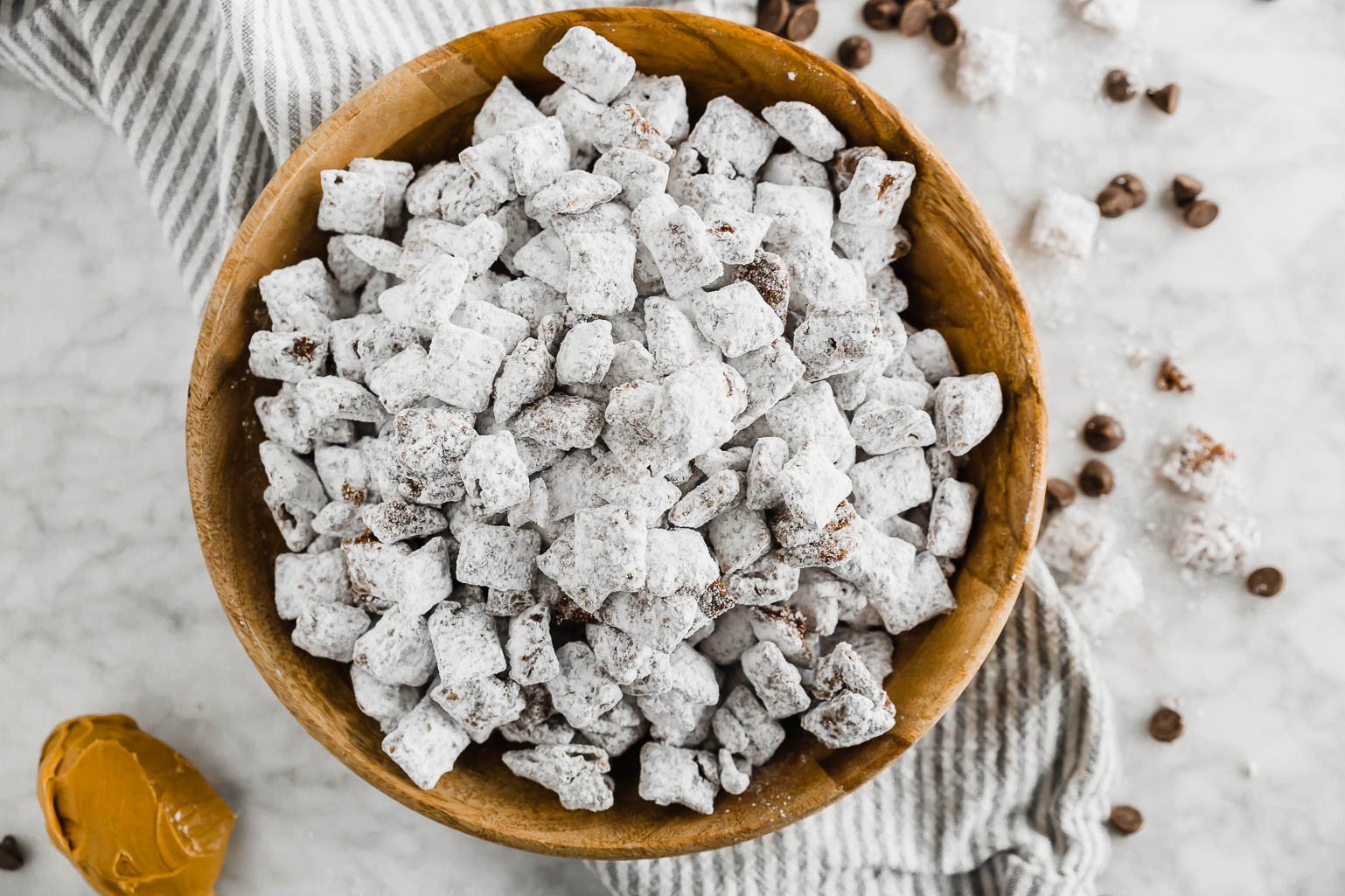 Dairy-free puppy chow in a wooden bowl with a spoonful of peanut butter and chocolate chips and powdered sugar on a linen napkin and table.