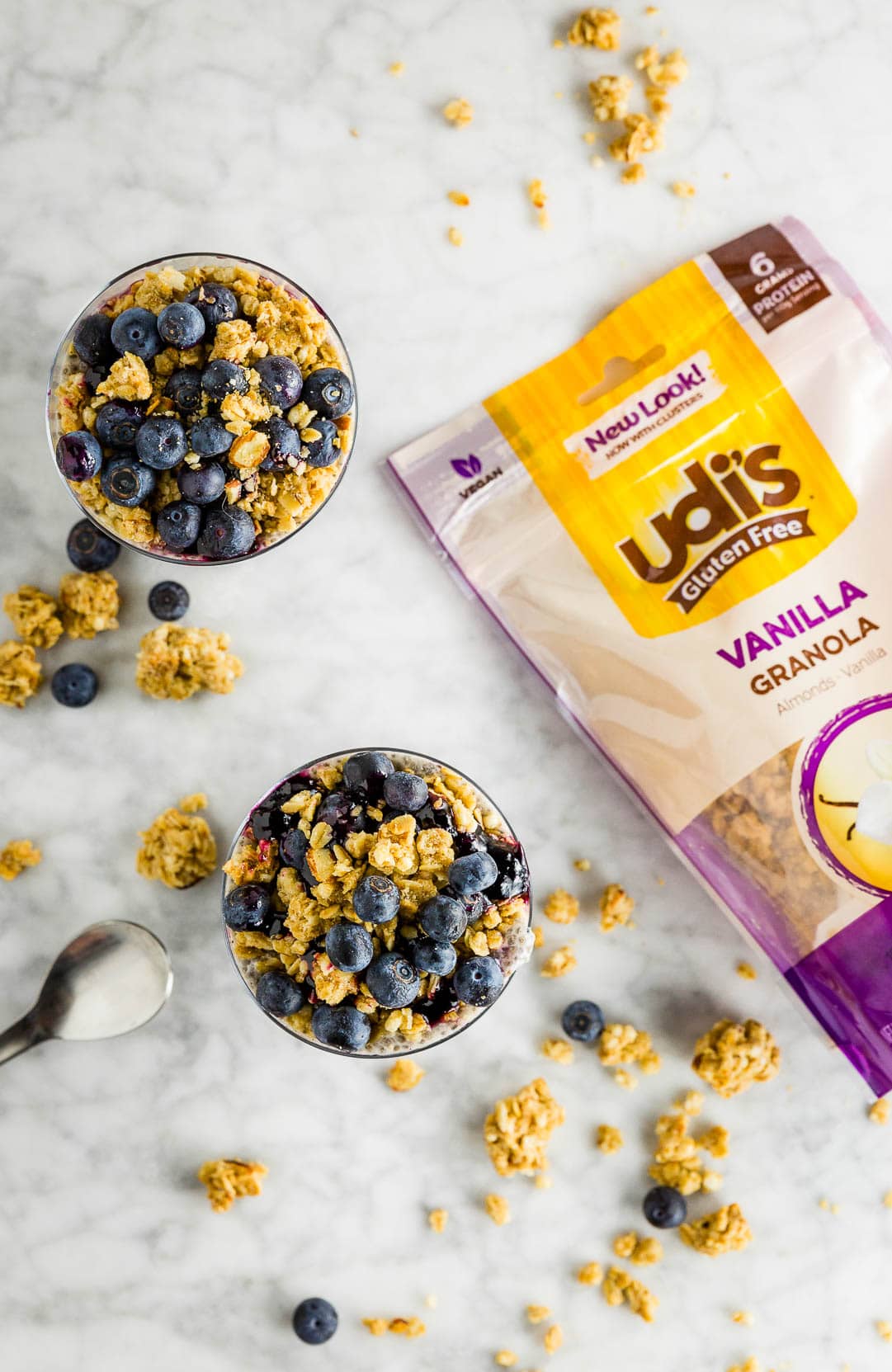 Two glasses filled with chia seed pudding with granola, blueberries and blueberry jam and a bag of Udi's Gluten Free Vanilla Granola on a table. 