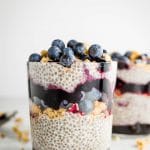 A glass filled with layered chia seed pudding with granola, jam and fresh blueberries.