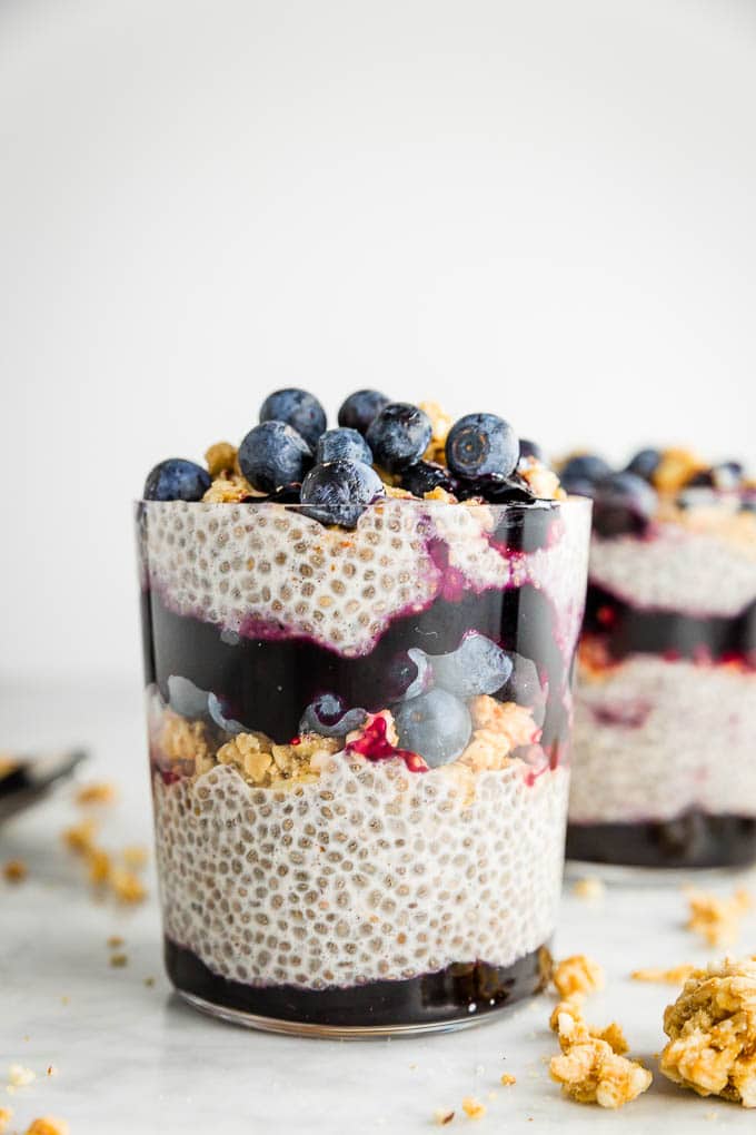 A glass filled with layered chia seed pudding with granola, jam and fresh blueberries.