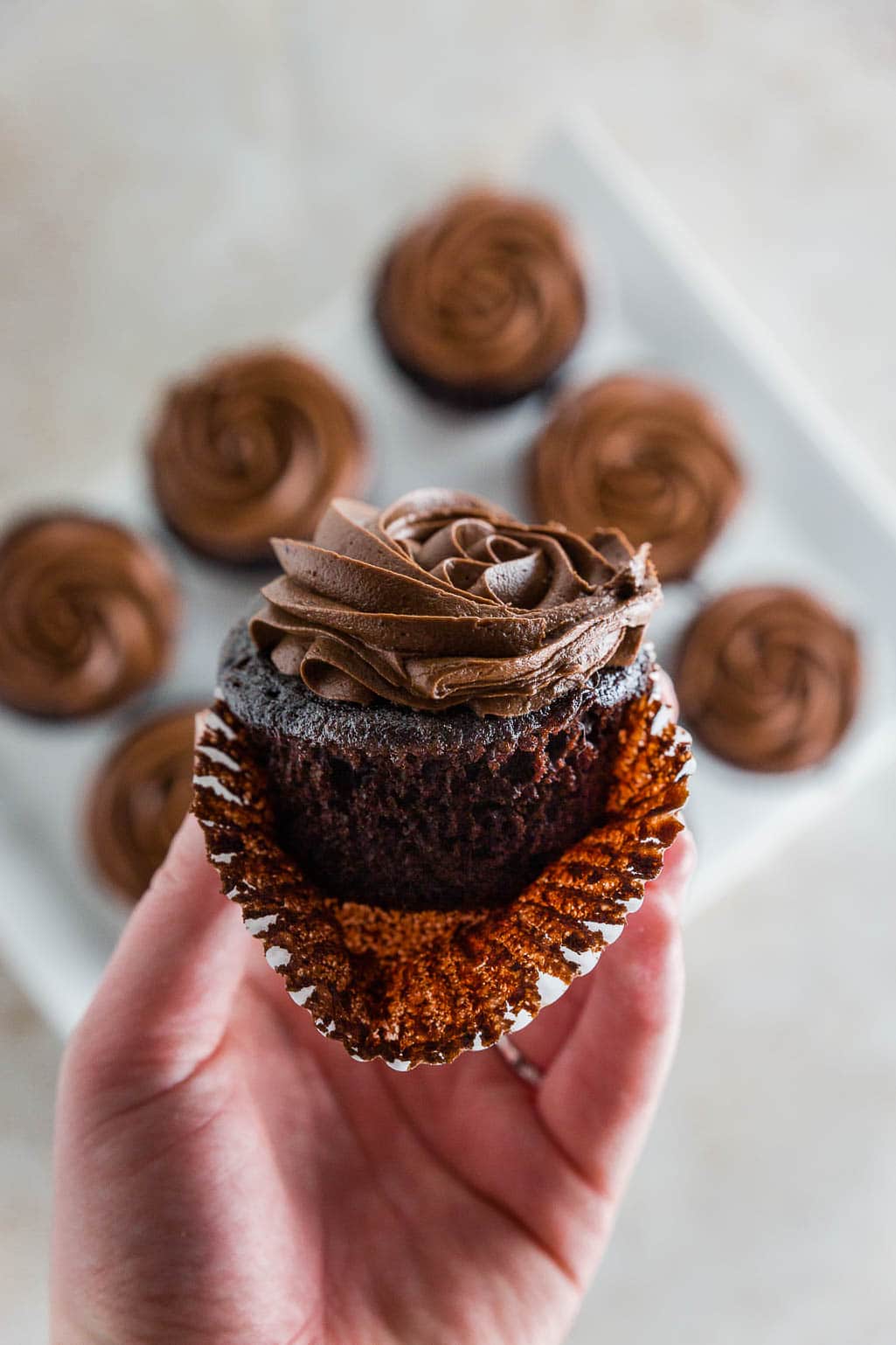 Photo of a hand holding a gluten-free chocolate cupcake with chocolate frosting with the cupcake liner peeled off. 