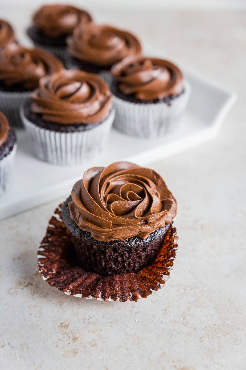 A gluten-free chocolate cupcake with swirled chocolate frosting sits in an open paper liner in the foreground, with more similarly decorated gluten-free chocolate cupcakes on a white tray in the background.