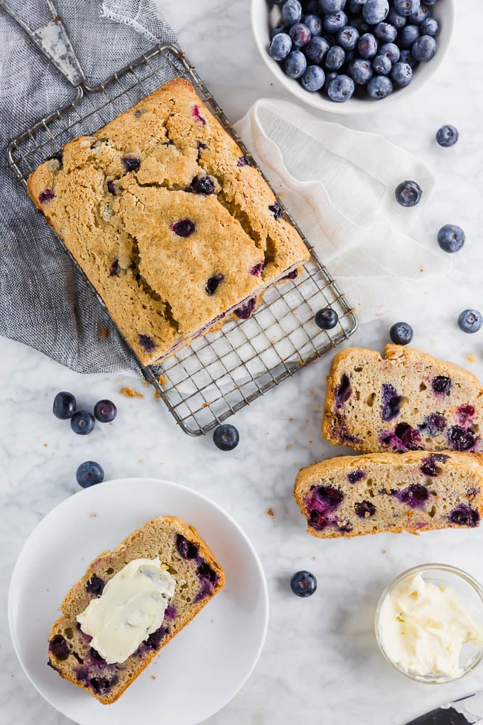 A loaf of gluten free blueberry muffin quick bread on a cooling rack with a slice on a plate with butter and a bowl of fresh blueberries. 