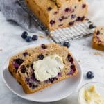 A loaf of blueberry muffin bread with two slices on a small plate slated in dairy free butter.