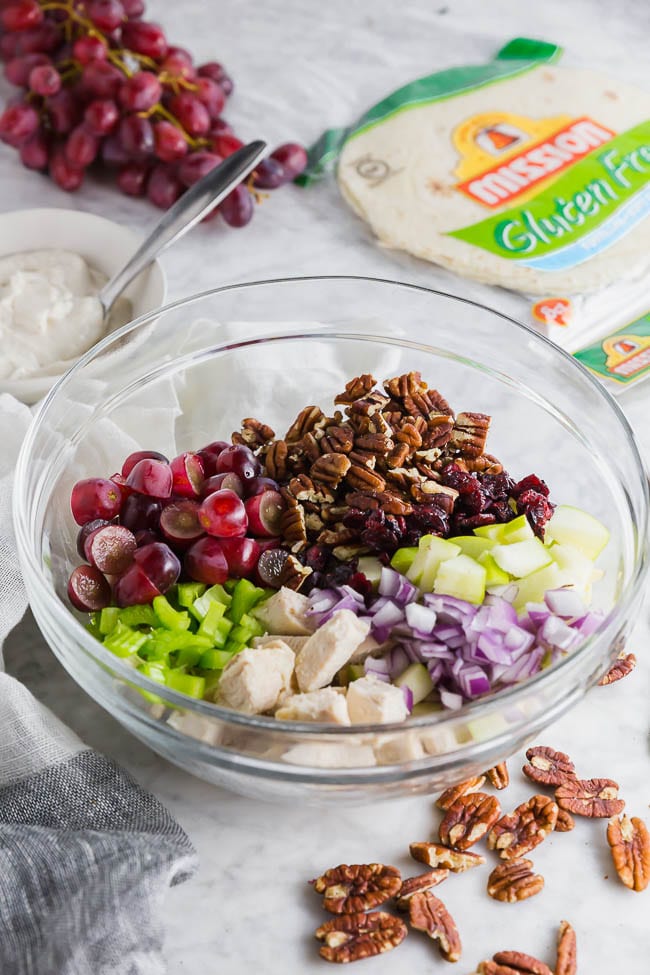 A photo of a large glass bowl containing grapes, pecans, cooked chicken, red onion, apples, celery and dried cranberries to make chicken salad. 