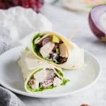 A photo of two gluten free chicken salad wraps on a plate with red onions and red grapes in the background.