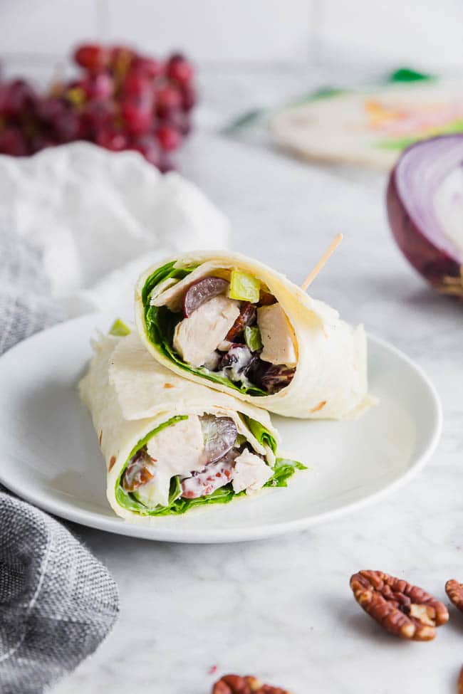 A photo of two gluten free chicken salad wraps on a plate with red onions and red grapes in the background. 