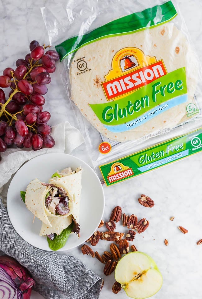A photo of two gluten free chicken salad wraps on a plate with a package of Mission Gluten Free tortillas and red grapes. 