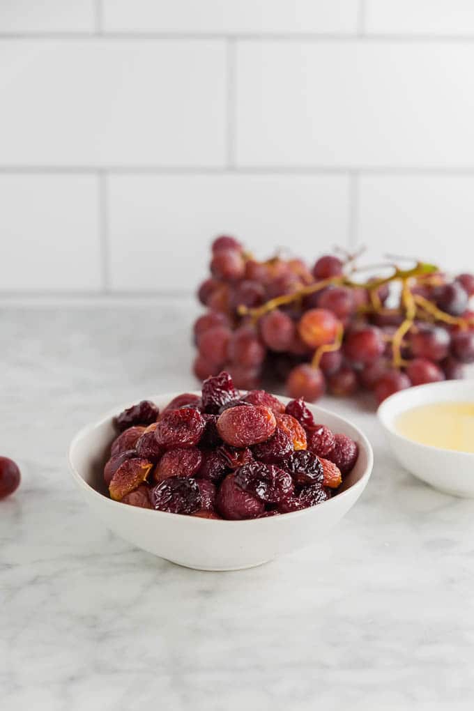 A bowl of roasted grapes on a table with fresh grapes and avocado oil.