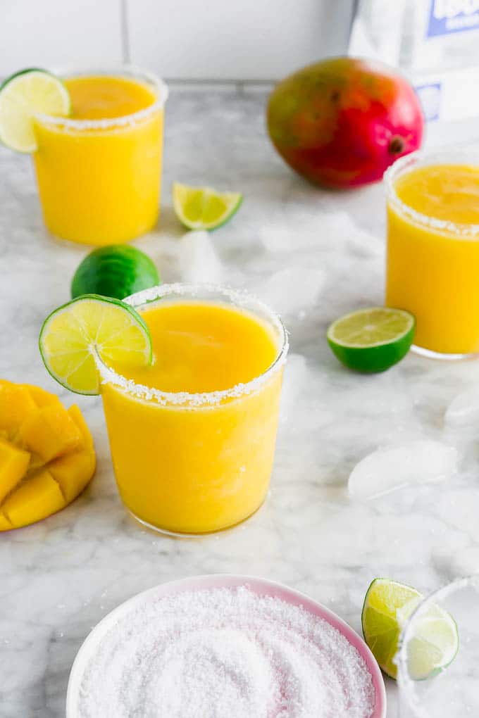 A photo of frozen mango margaritas, a plate with kosher salt, limes, fresh mangos and a bottle of tequila. 