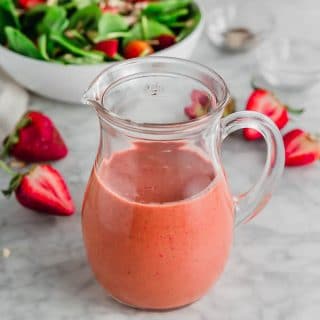 A photo of a glass pitcher with strawberry balsamic vinaigrette with fresh sliced strawberries and a spinach salad in the background.