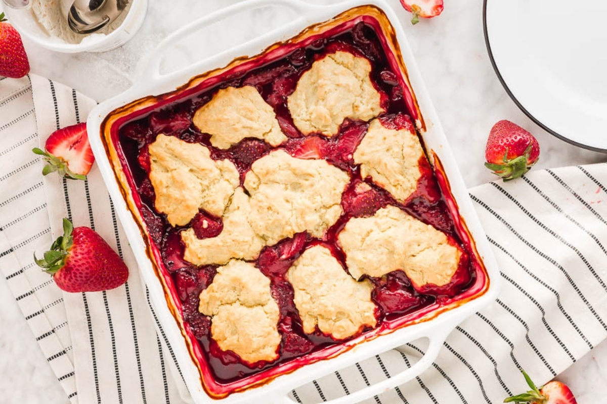 A square white baking dish filled with gluten-free strawberry cobbler, topped with golden-brown biscuit pieces. Fresh strawberries are scattered around on a striped cloth beside a white plate and spoon.