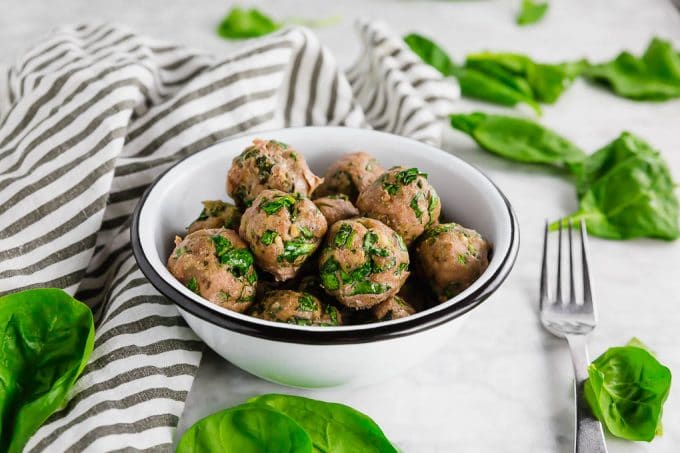 A bowl of gluten free turkey meatballs with spinach on a white table with fresh spinach leaves and a fork ready to dig in.