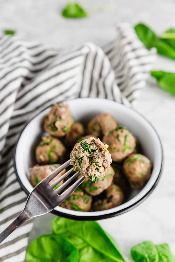 A bowl of baked turkey spinach meatballs with one meatball on a fork showing the inside of the gluten free turkey meatball. 