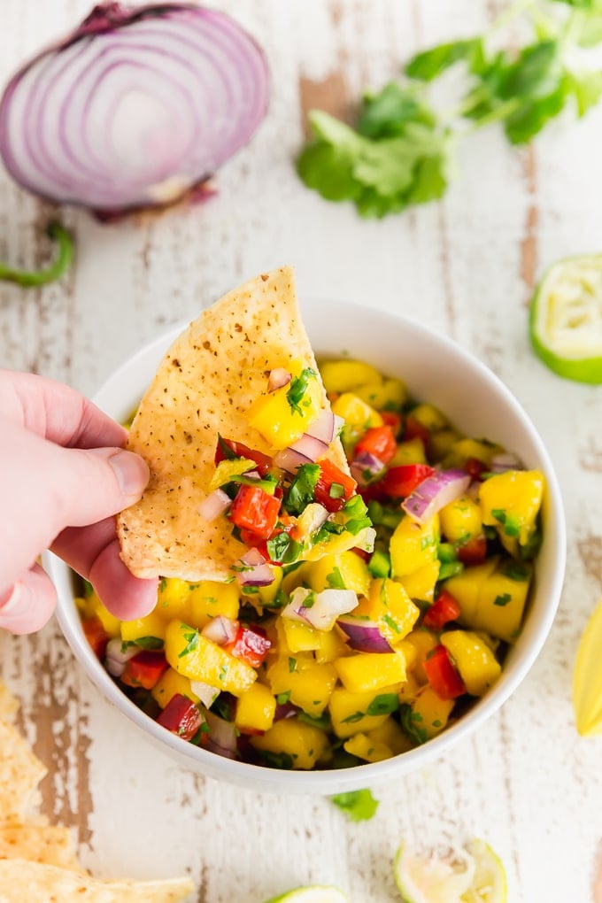 A photo of a hand holding a chip topped with mango bell pepper salsa over a bowl of fruit salsa. 