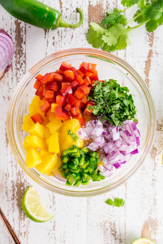 A bowl of ingredients for mango salsa including cilantro, red bell pepper, red onion, jalapeÃ±o, mango, salt and lime juice.
