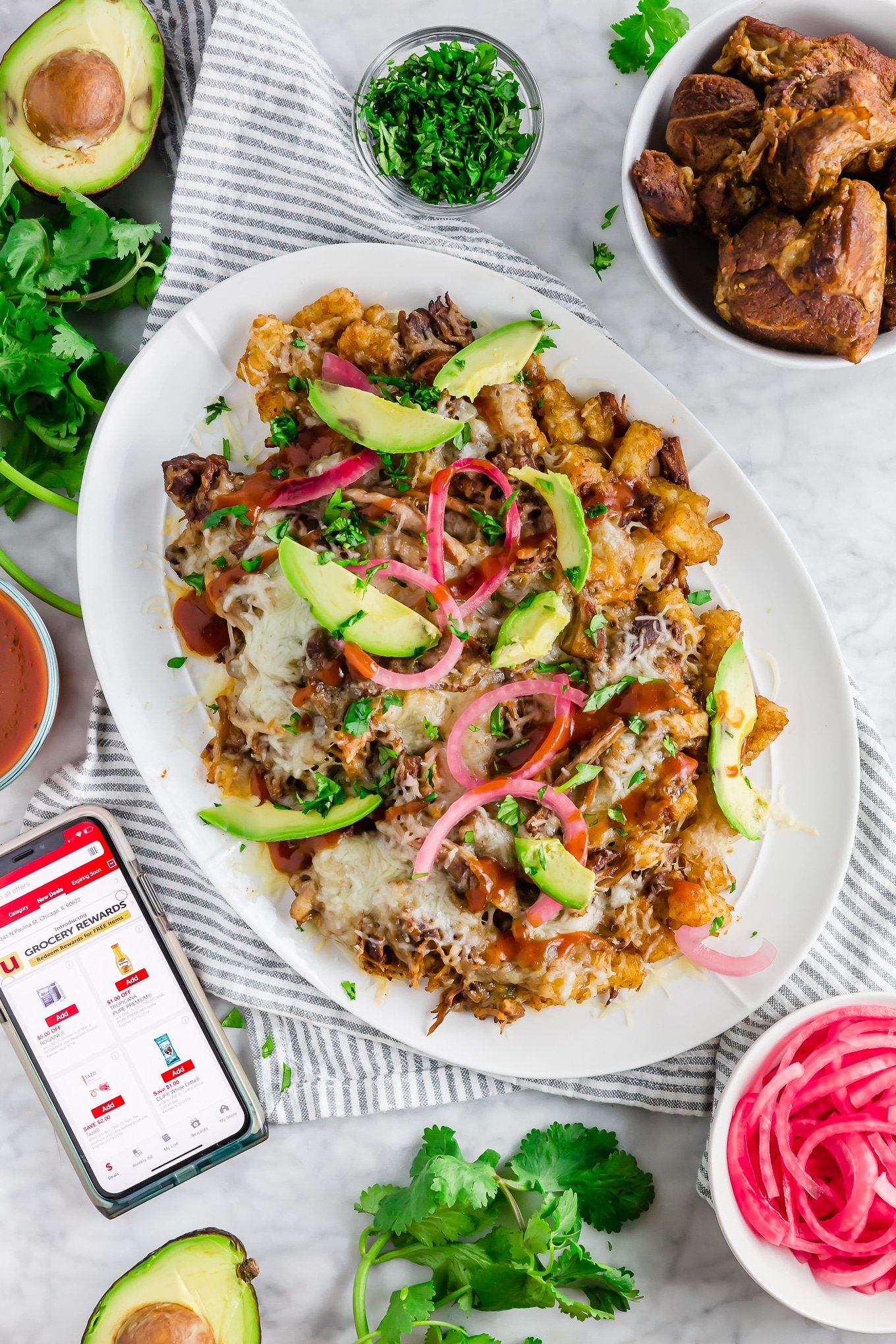 A photo of a platter of BBQ Pulled Pork Tater Tots topped with pickled onions, avocado and cilantro. 