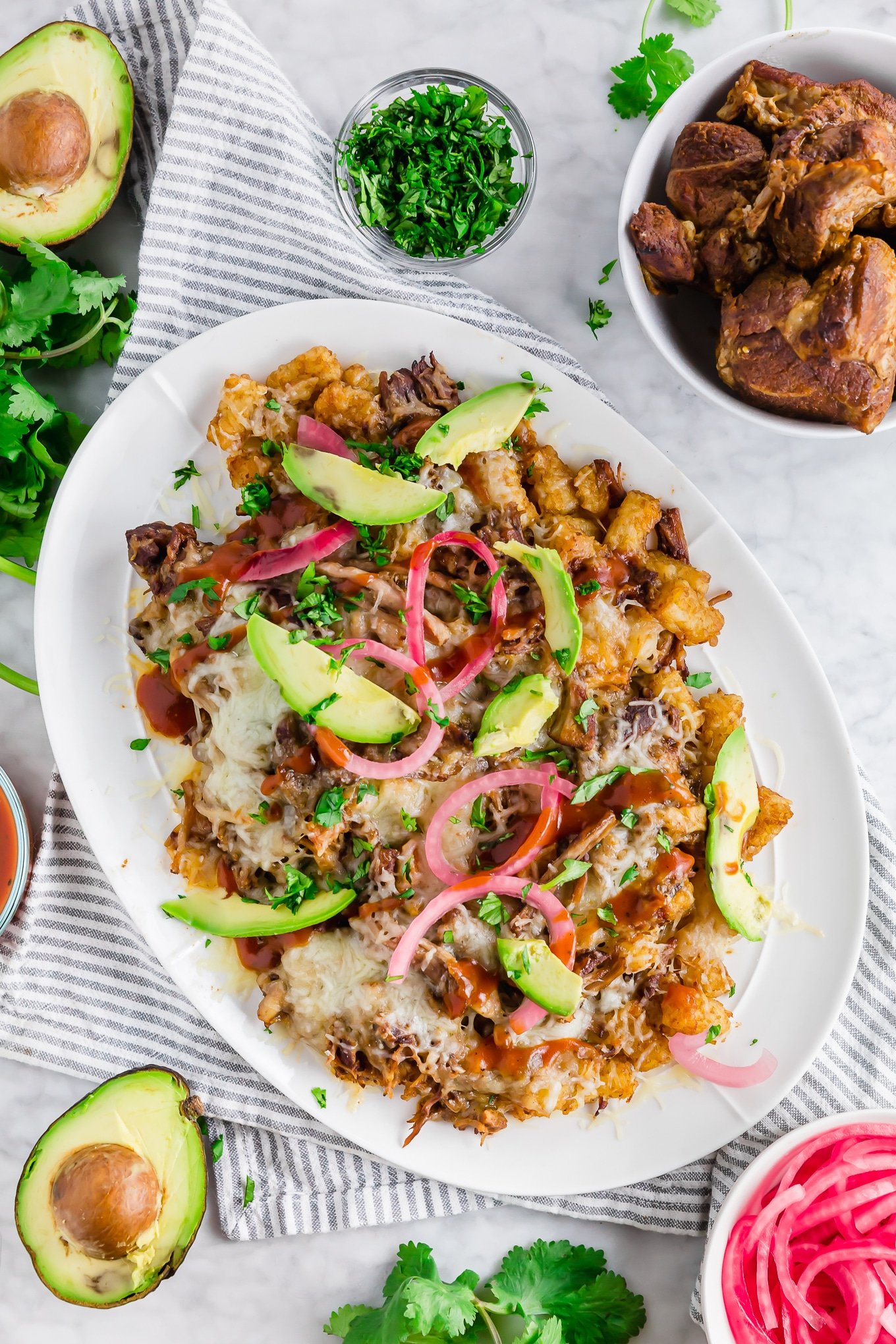 A photo of a platter of loaded tater tots topped with BBQ pulled pork, pickled onions, shredded cheddar cheese, pickled onions and cilantro. 