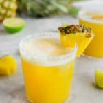 A photo of a glass filled with pineapple margarita with a wedge of fresh pineapple and lime.