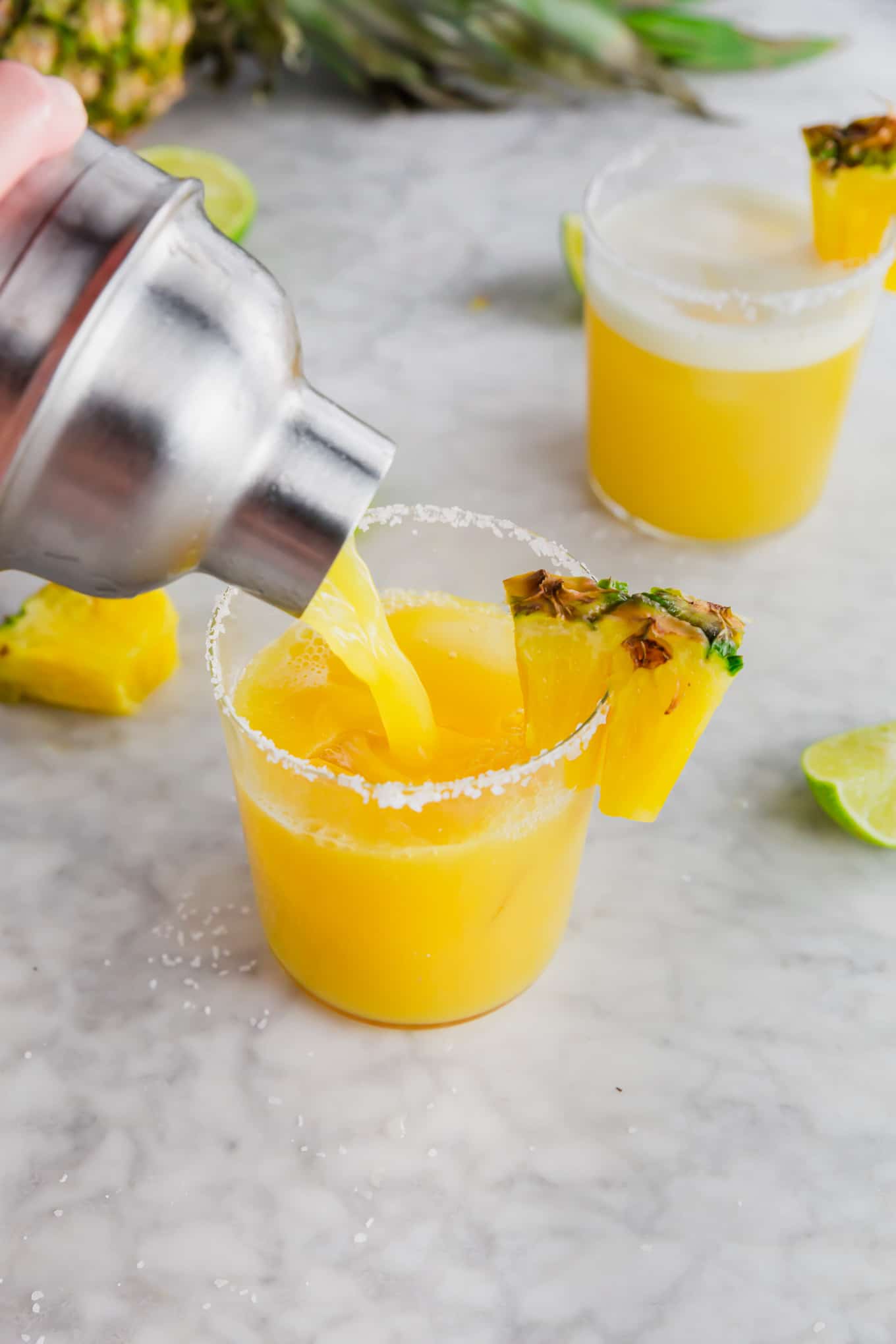 A cocktail shaker pouring out pineapple margarita into a glass rimmed with kosher salt with a pineapple wedge.