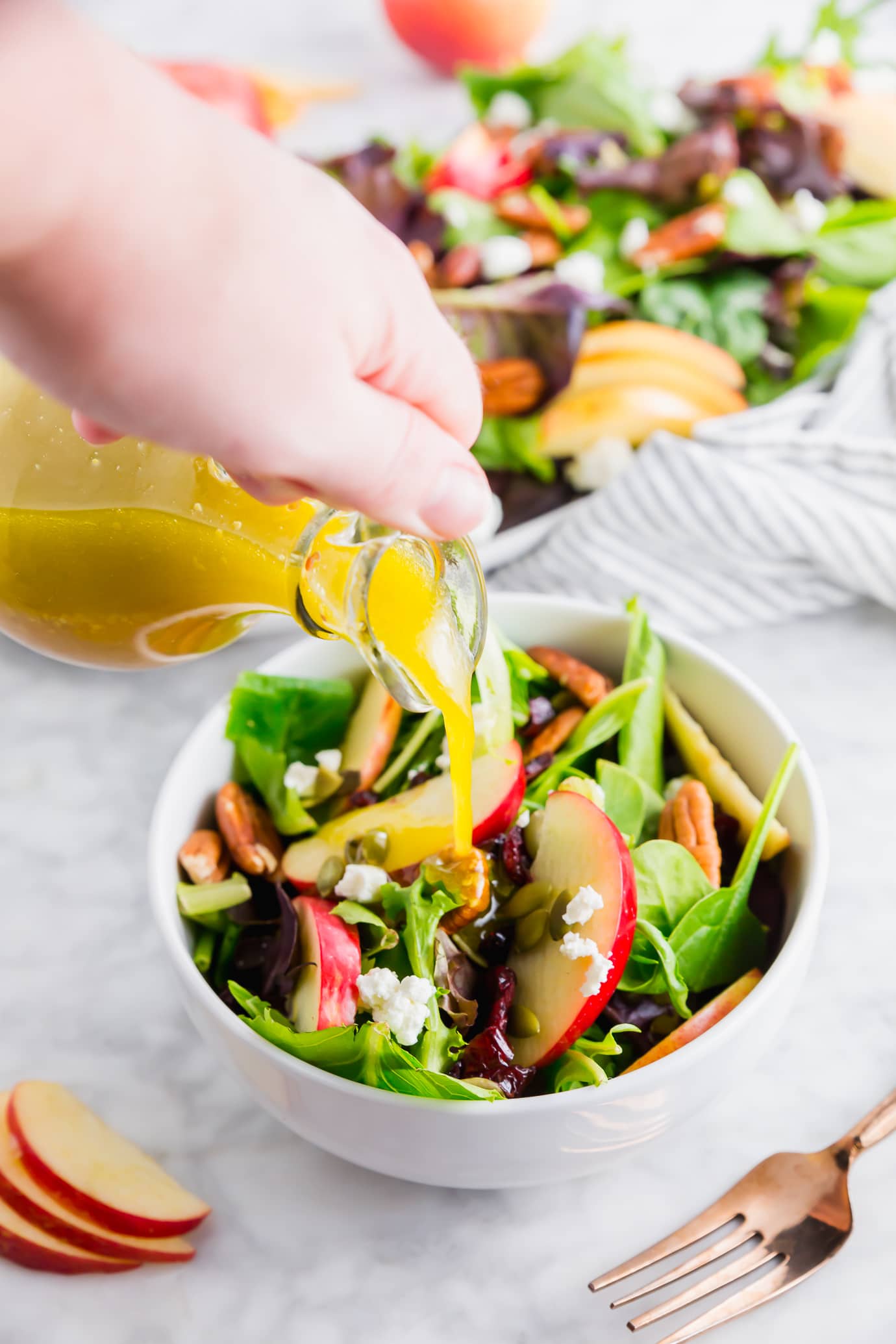 A photo of a spinach and apple salad with a bottle of homemade apple cider vinaigrette being poured over the top.